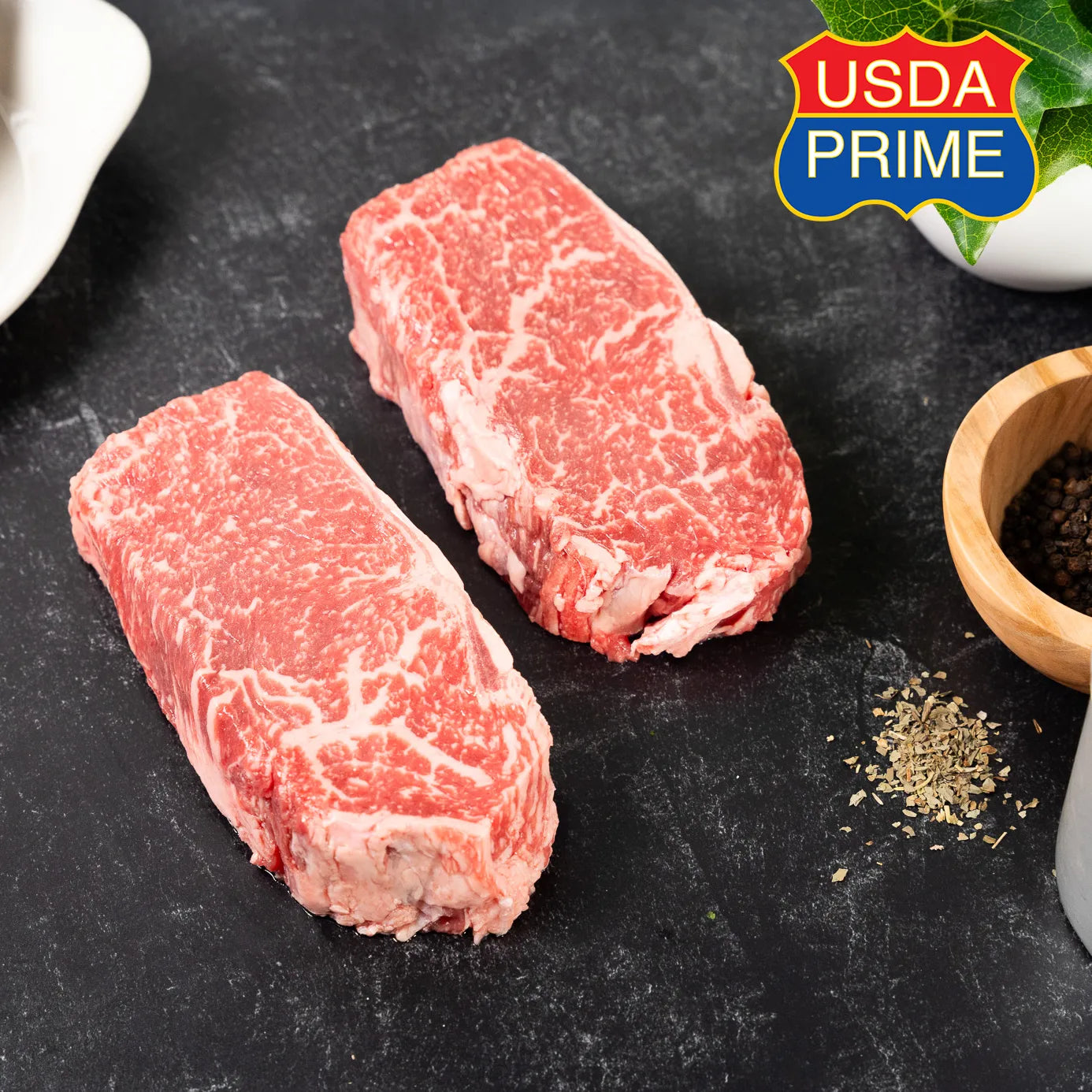 Two raw USDA Prime Heritage Grass-Fed New York Strip steaks on a dark slate surface, showing rich marbling throughout. A USDA Prime badge appears in the corner, with peppercorns and herbs nearby.