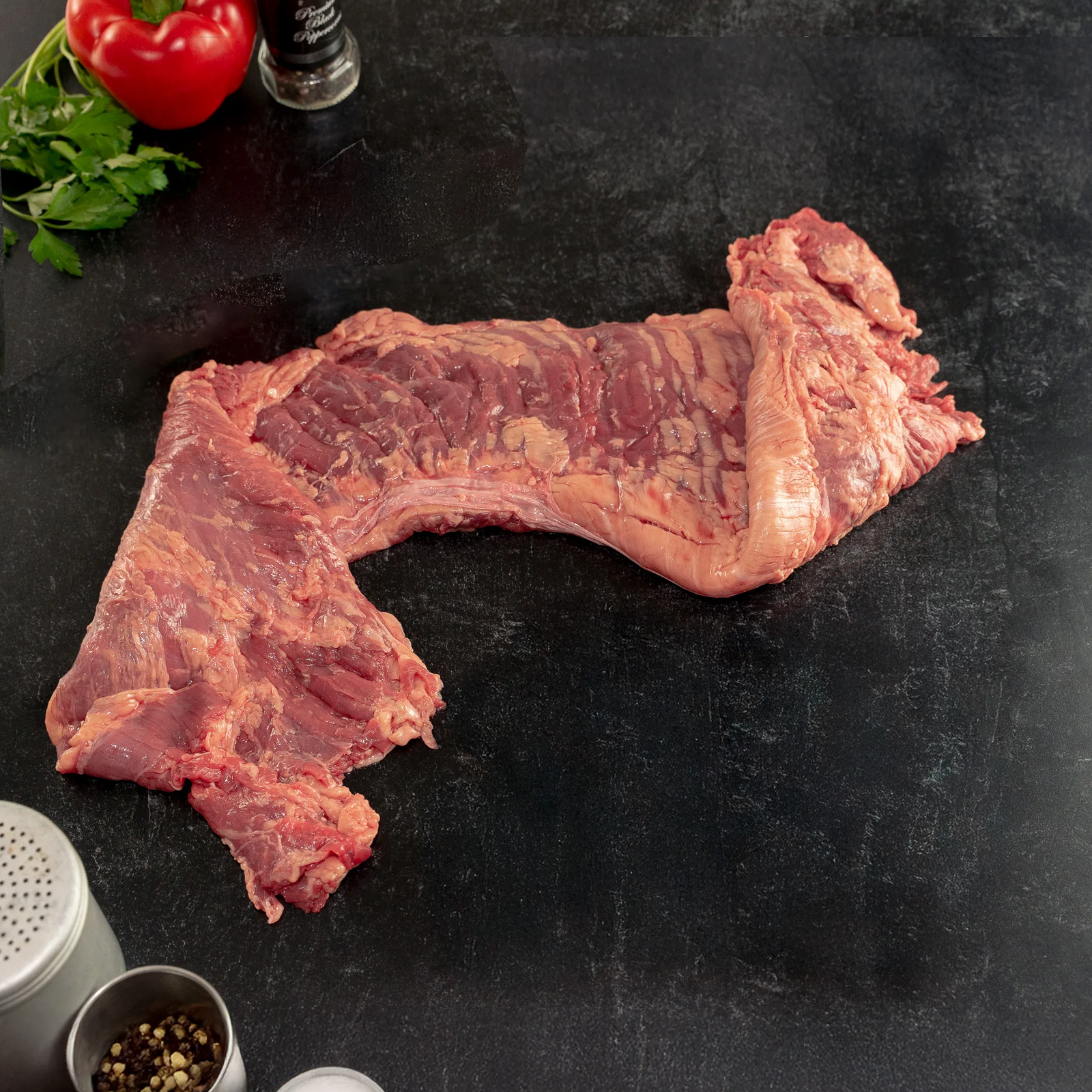 Raw grass-fed outside skirt steak from Joyce Farms’ Heritage Aberdeen Angus line, shown on a dark prep surface with visible marbling and natural texture, surrounded by herbs, peppercorns, and seasoning tins.