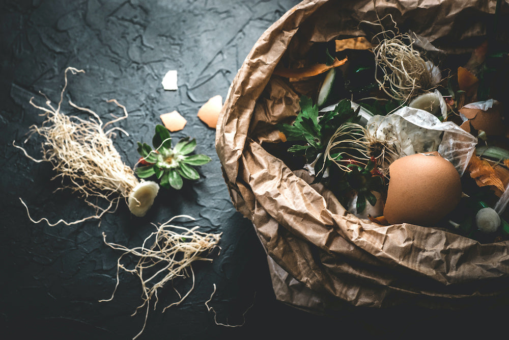 GPT A compost pile inside a brown paper bag is set against a dark, textured background. Organic waste such as eggshells, tea bags, carrot peelings, and wilted herbs with their roots still attached spill slightly out of the bag. 