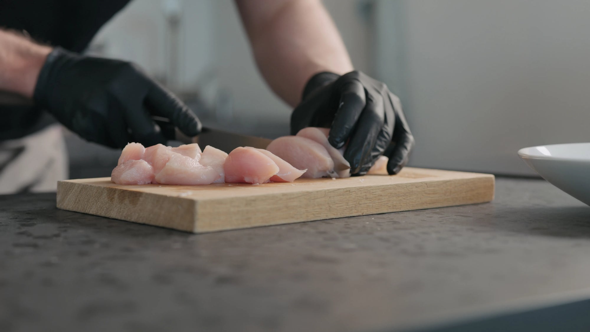 Person wearing black gloves cutting raw chicken breasts on a wooden cutting board.