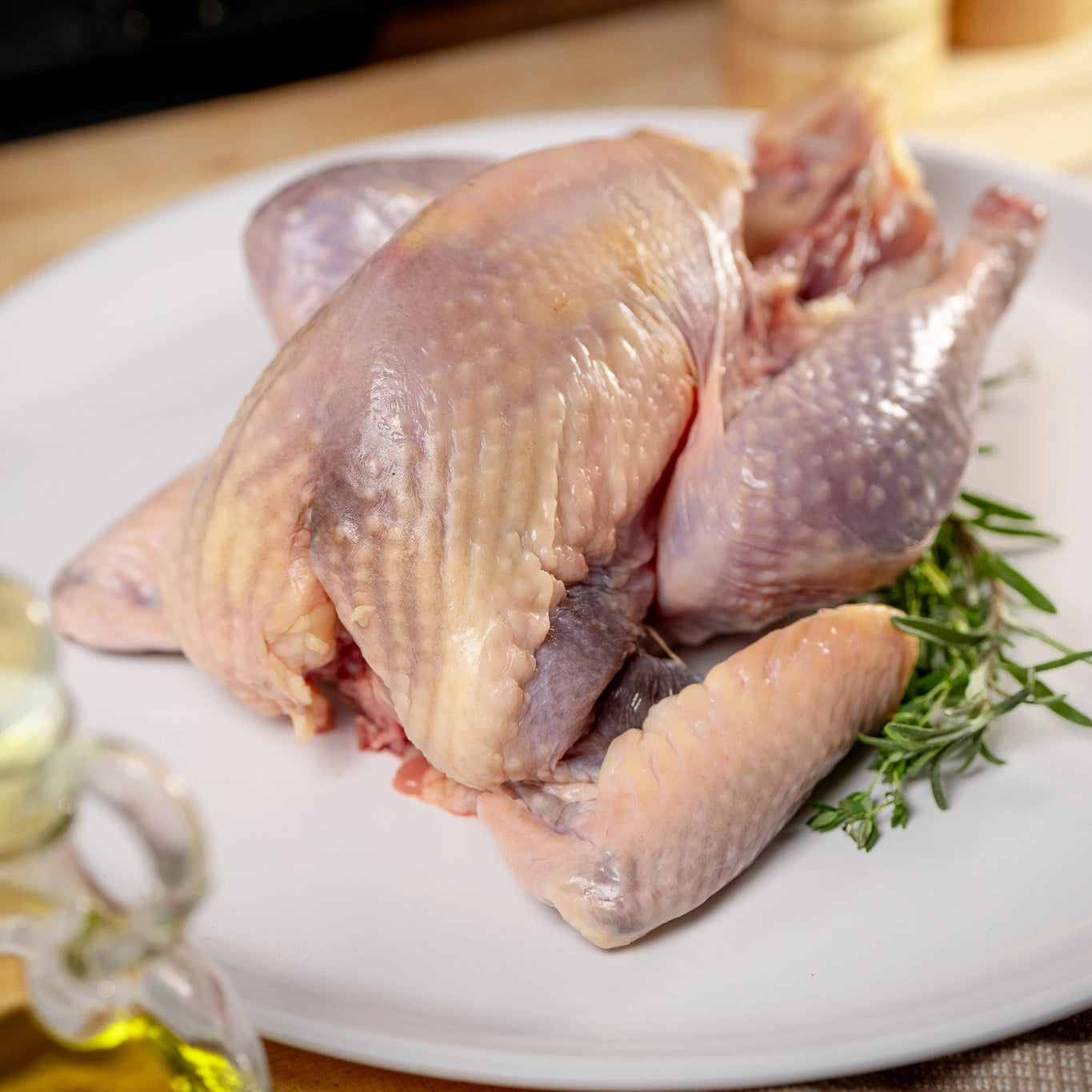 A raw Joyce Farms Heritage Pintade (French Guinea) trussed and ready for roasting, displayed on a white plate with rosemary and thyme on the side