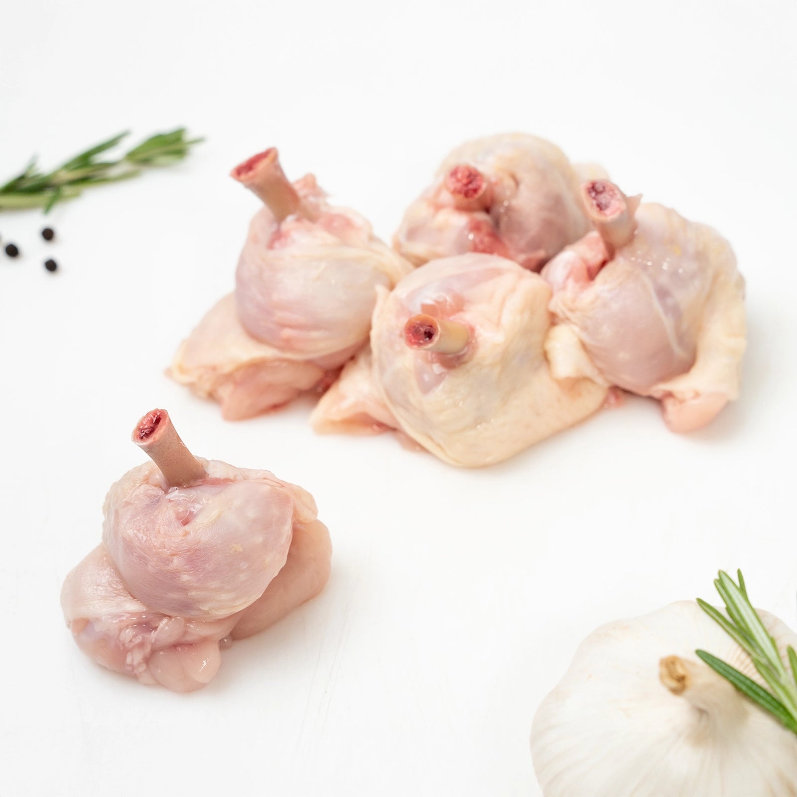 Five raw Joyce Farms Naked Chicken Frenched Drumettes arranged together on a white cutting board with their frenched bone tips facing upwards. Garnish of rosemary, peppercorn, and garlic rest on the side.
