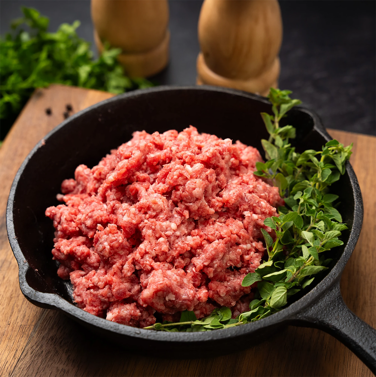 Joyce Farms Signature Angus Ground Beef raw in a black cast iron skillet with herbs on a wooden surface