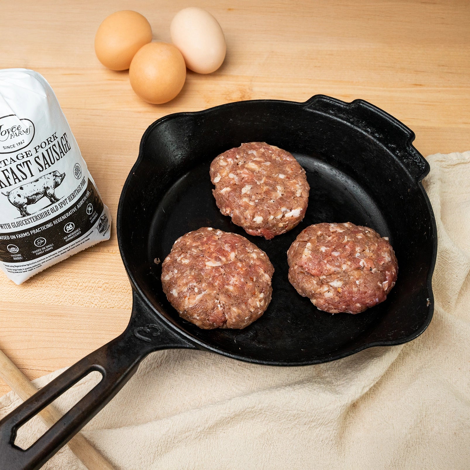 Joyce Farms Heritage Pork Breakfast Sausage in the form of three patties sit in a cast iron pan. The pork sausage product bag sits next to it.