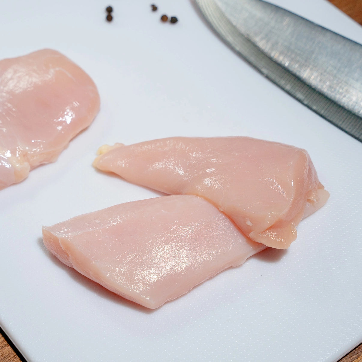 Raw Antibiotic-Free Naked Chicken Breast Medallions resting on a white cutting board with peppercorn and a kitchen knife resting on the side.