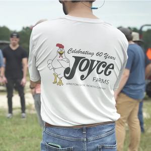 Person wearing a Joyce Farms Limited Edition 60th Anniversary T-Shirt at an outdoor farm event, featuring a cartoon chicken and the text 'Celebrating 60 Years Joyce Farms, Winston-Salem, North Carolina' on the back.