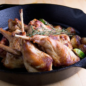 beautifully prepared dish of Joyce Farms Naked Rabbit, cooked to a golden brown in a cast iron skillet. The rabbit pieces are arranged with their tender, caramelized surfaces facing upward, showcasing a rich, savory glaze. Fresh sprigs of thyme are placed on top, adding an aromatic touch to the dish. Surrounding the rabbit are roasted baby potatoes, slightly charred and glistening, complementing the rustic and hearty presentation.