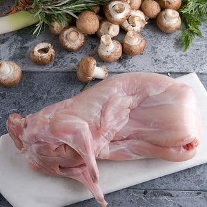 raw, whole Joyce Farms Naked Rabbit placed on a marble cutting board. The rabbit's pale, pinkish flesh is clean and neatly prepared, ready for cooking. Surrounding the rabbit are fresh mushrooms, herbs like rosemary, and possibly leeks, creating a rustic, culinary setting. The combination of ingredients suggests preparation for a classic, hearty dish.