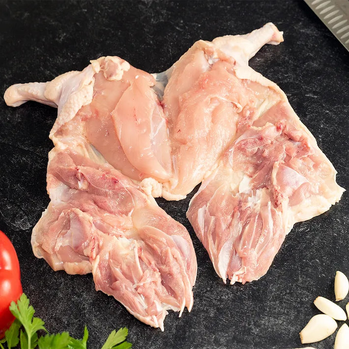 The reverse side of a raw, semi-boneless Poulet Rouge Heritage Chicken, prepared on a dark slate surface, ready for cooking, surrounded by fresh garlic cloves and parsley.