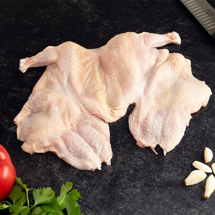 A raw, semi-boneless Poulet Rouge Heritage Chicken laid flat on a dark slate surface, with skin intact, accompanied by fresh garlic cloves and parsley.