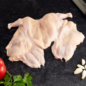 A raw, semi-boneless Poulet Rouge Heritage Chicken laid flat on a dark slate surface, with skin intact, accompanied by fresh garlic cloves and parsley.