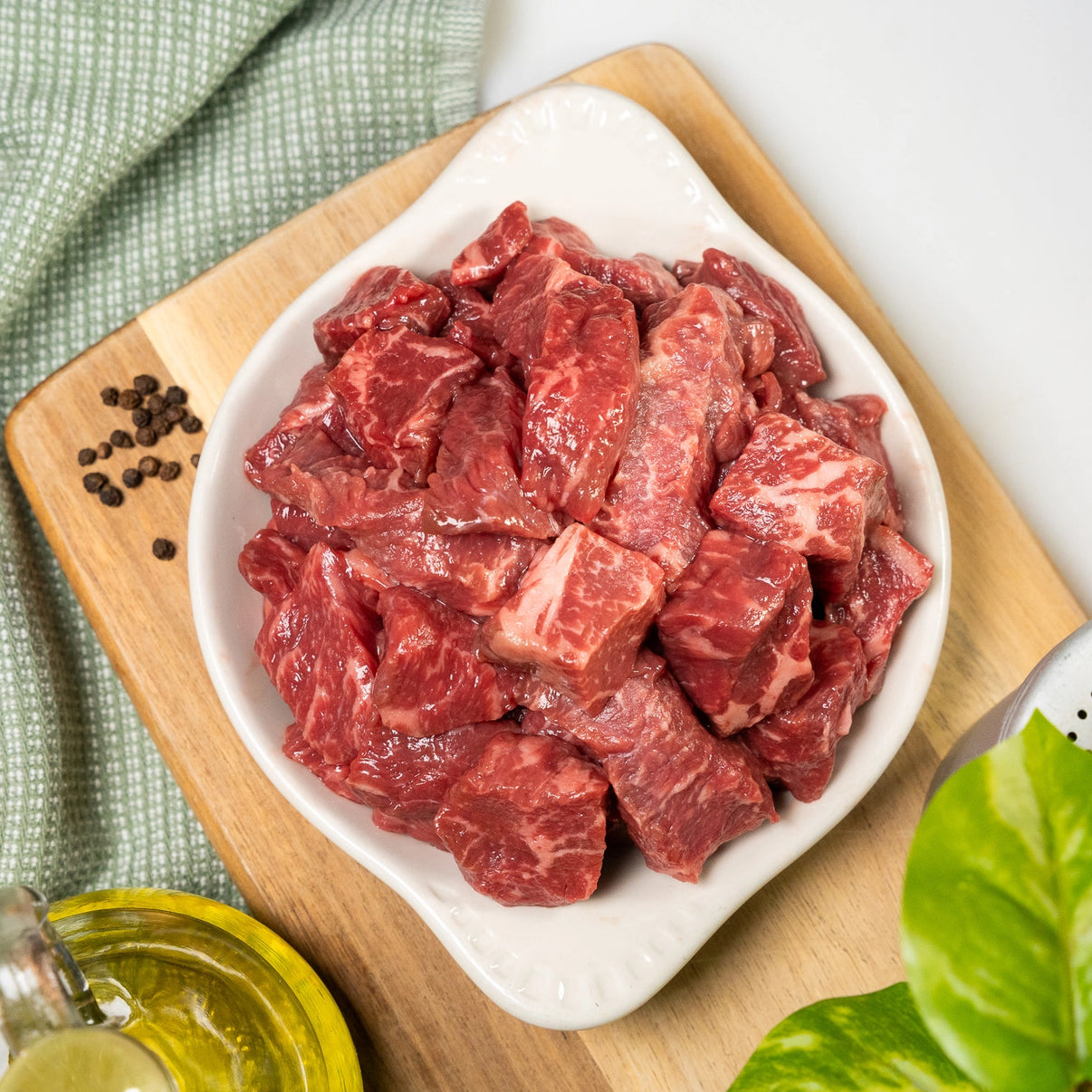 Raw beef chunks in a white bowl on a wooden cutting board with a green cloth and plant in the background.