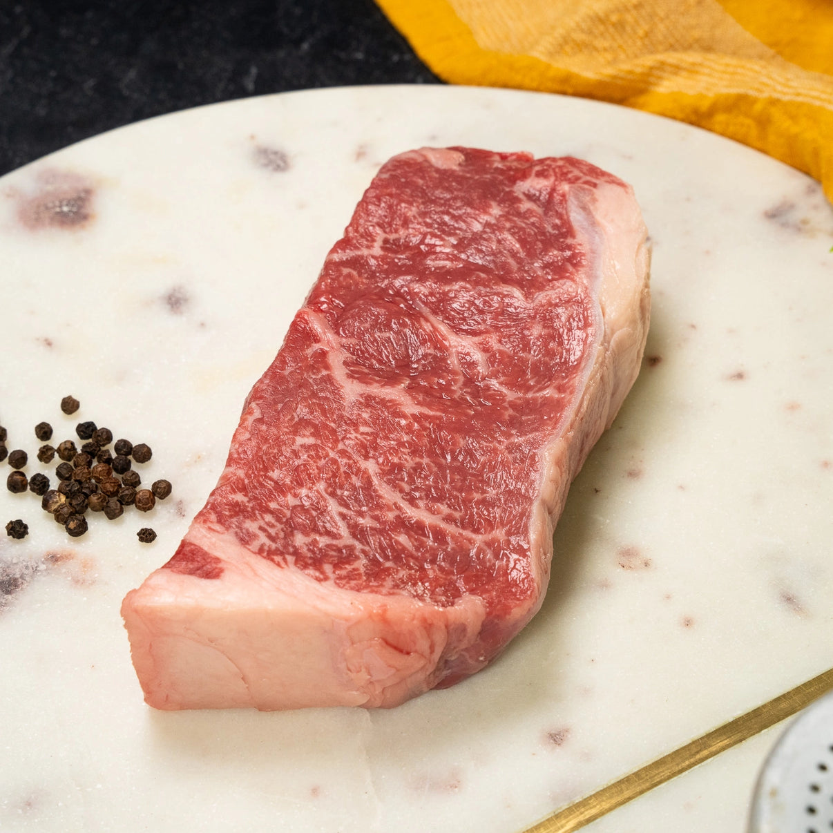 A raw Joyce Farms New York Strip Steak USDA Grade Prime with fat marbling rests on a white marble plate with peppercorns sprinkled on the side and a yellow napkin in the background.