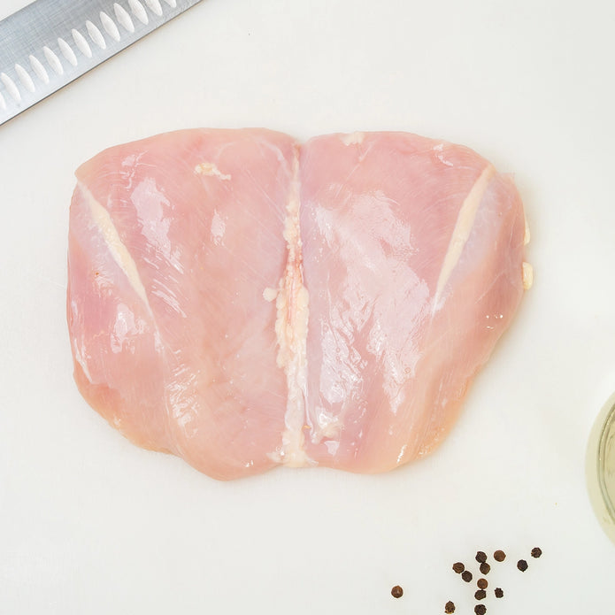 Raw Joyce Farms Boneless, Skinless Naked Chicken breast on a cutting board with a knife and peppercorns.