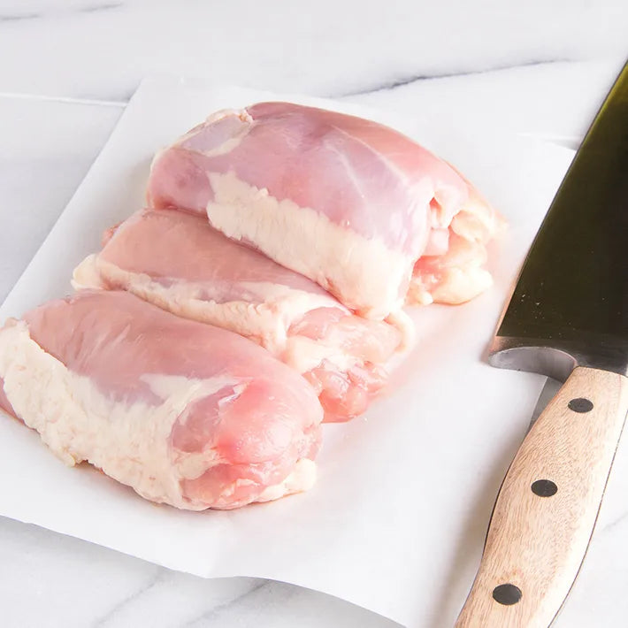 Raw antibiotic-free Naked Chicken Bone-In, Skin-On Chicken Thighs on a white surface with a knife.
