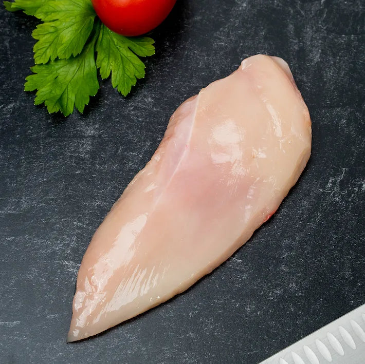 Raw Poulet Rouge Boneless Skinless Chicken Breast from Joyce Farms, a heritage breed known for its slow-growing, pasture-raised, air-chilled, and antibiotic-free process, displayed on a black cutting board with a tomato and parsley garnish. 