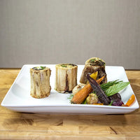 Three pieces of Joyce Farms Heritage Grass-Fed Beef Bones, roasted and served on a white plate with a colorful assortment of roasted vegetables.