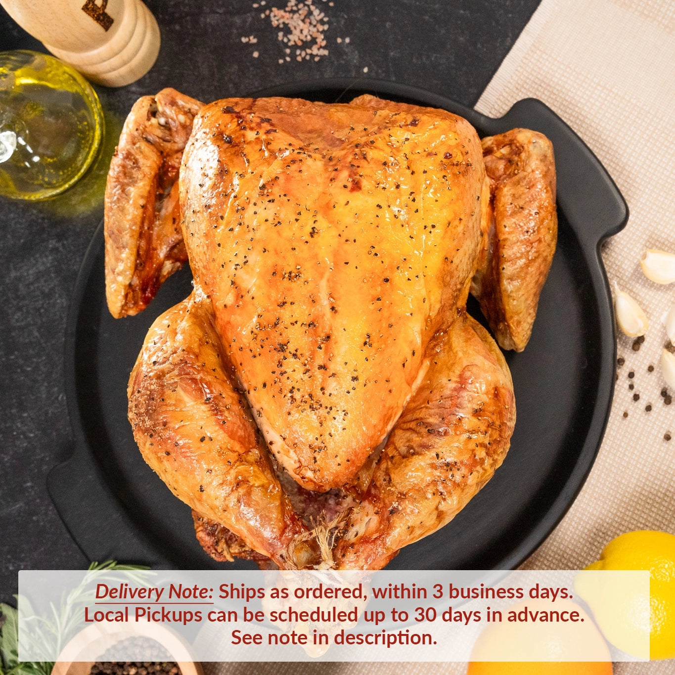 A whole roasted turkey on a black serving platter, seasoned with salt and pepper, surrounded by lemons, garlic cloves, fresh herbs, and a bottle of oil on a dark tabletop. Text overlay reads: “Delivery Note: Ships as ordered, within 3 business days. Local Pickups can be scheduled up to 30 days in advance. See note in description.