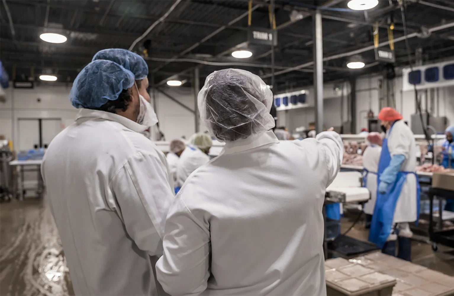 Joyce Farms employees working in a processing facility, dressed in white lab coats, hair nets, and masks. One employee is pointing and giving instructions to another, highlighting a collaborative and organized work environment.