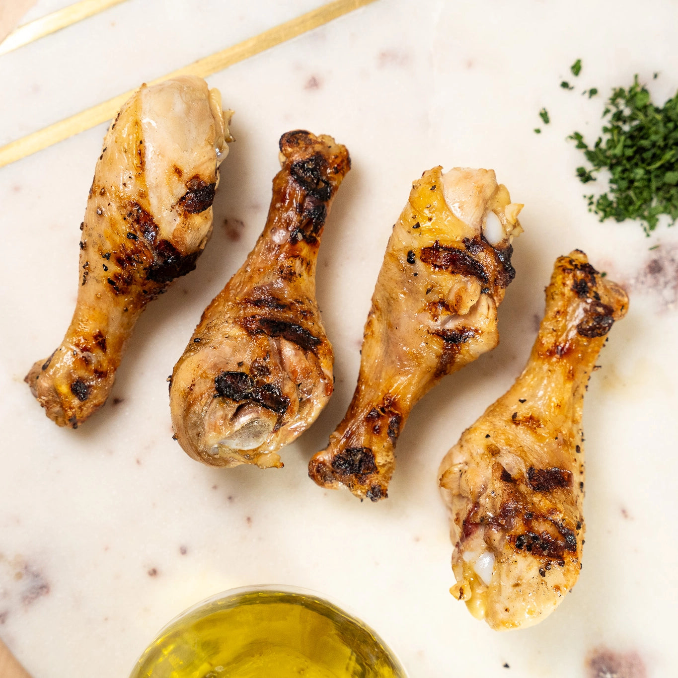 Grilled chicken drumsticks on a marble surface with a small bowl of oil and herbs.
