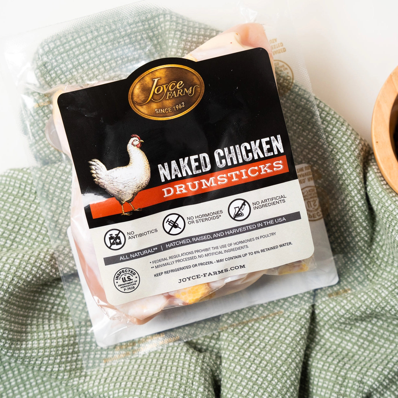 An individual package of Joyce Farms Naked Chicken Drumsticks resting on a green kitchen towel