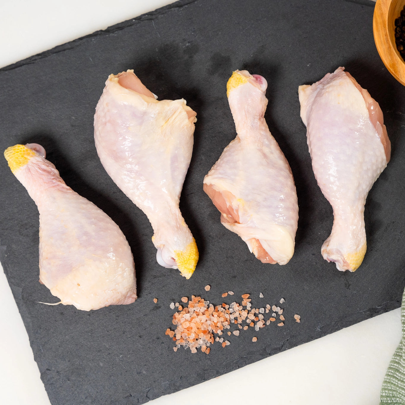 Raw chicken drumsticks on a black slate surface with salt and pepper.
