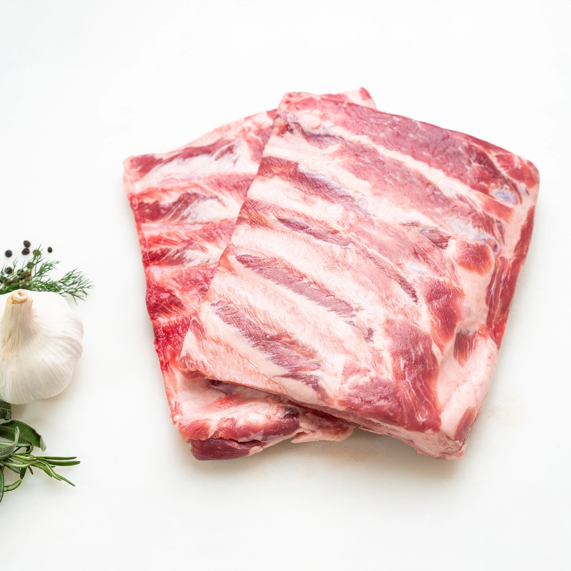 Two racks of Joyce Farms Heritage Pork Spare Ribs rest on top of one another on a white cutting board, displaying the marbling and texture of the meat. Garlic, peppercorn, and other green garnishes rest next to it ready for prepping.