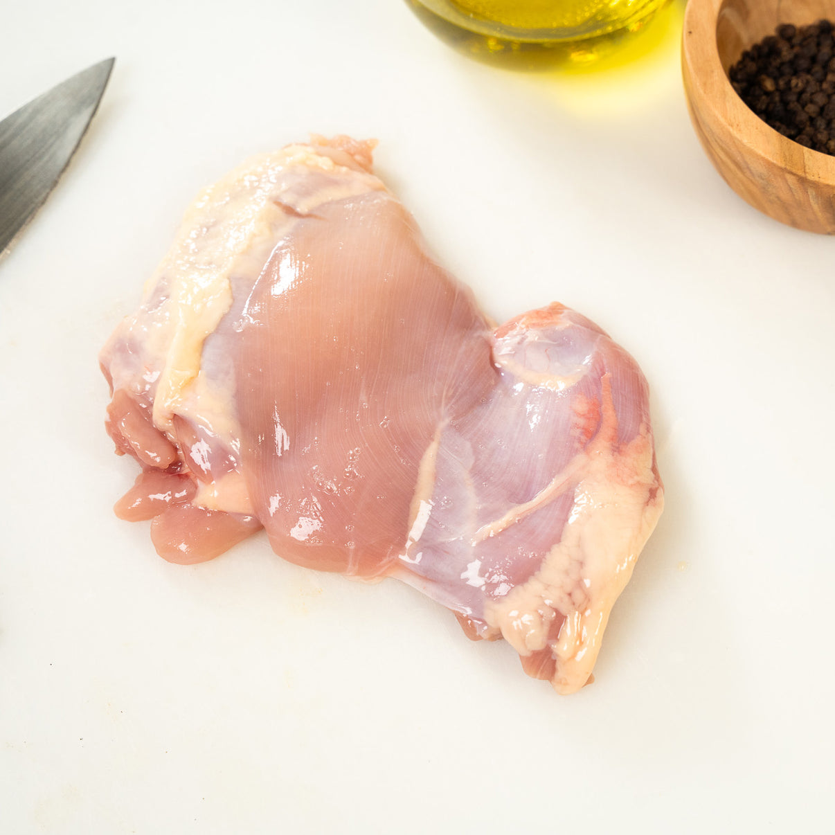 Raw joyce farms boneless, skinless chicken thigh on a cutting board with oil and peppercorns in the background