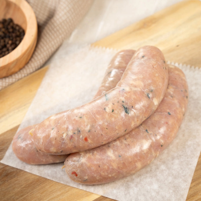Joyce Farms Raw Naked Mild Italian Chicken Sausages stacked on top of wax paper, resting on a wooden cutting board with a small wooden ramekin with peppercorn inside, resting on tan hand towel.