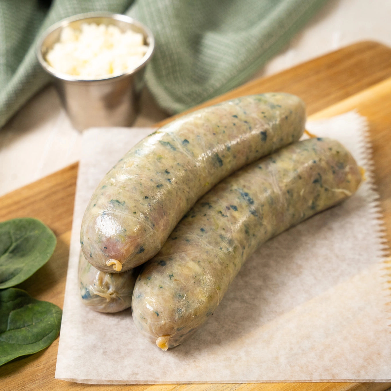 Joyce Farms, all-natural, no antibiotic or growth hormone, raw naked chicken spinach and feta sausage links stacked on wax paper that rests on a wooden cutting board. Spinach leaves, a ramekin of feta, and a green hand-towel rest nearby.