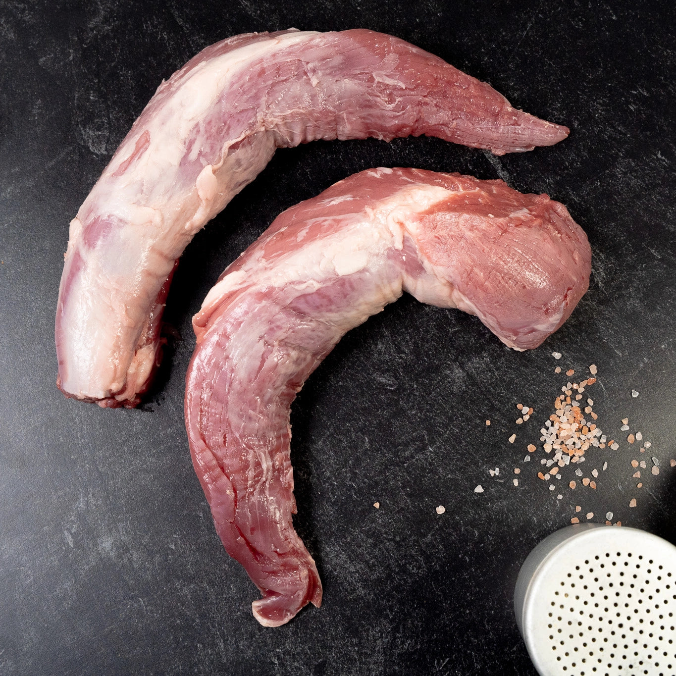Two raw Joyce Farms Heritage Pork Tenderloin pieces on a black surface with coarse salt and a seasoning shaker.