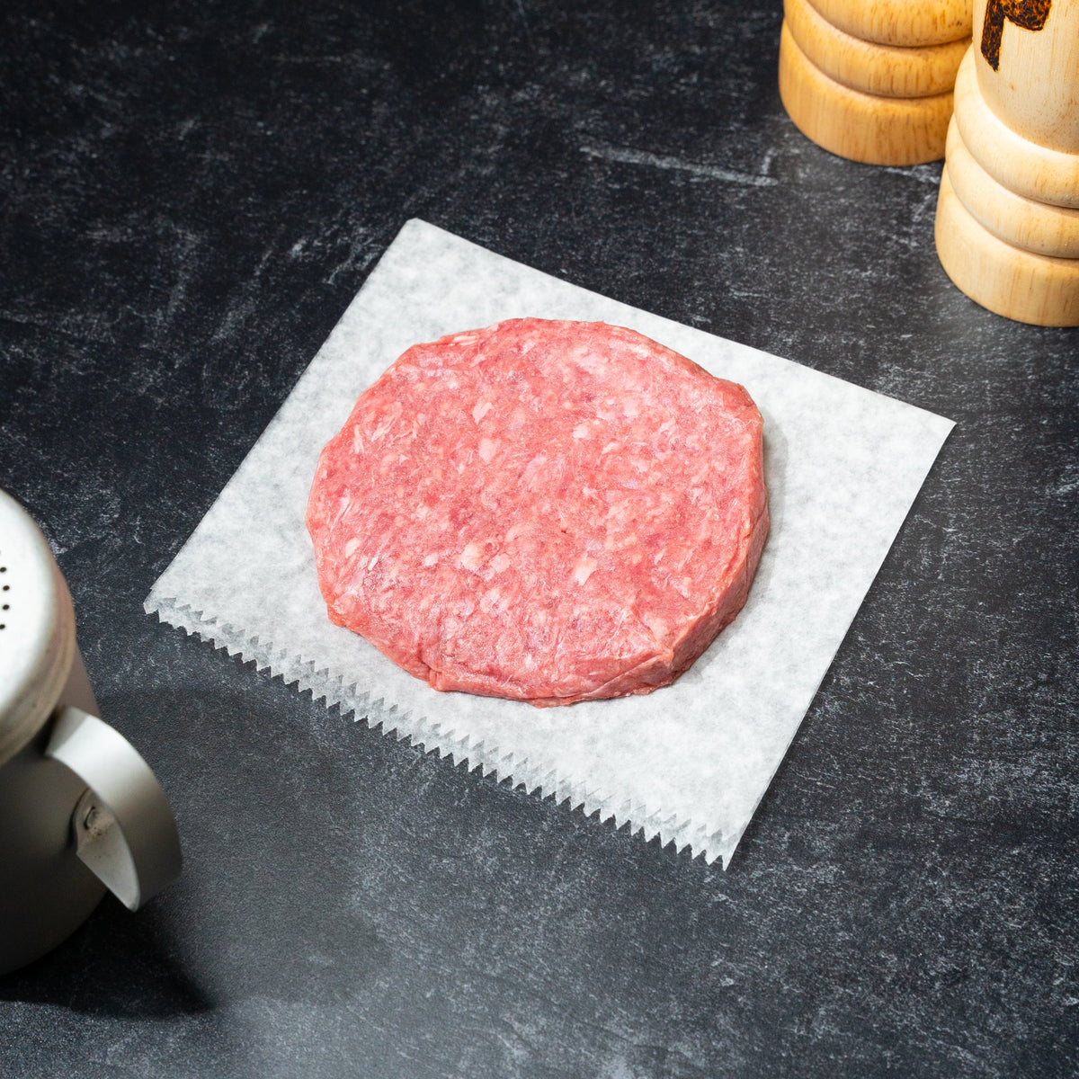 A single round raw beef patty sits on a square sheet of white wax paper on a dark textured countertop, with partial kitchen props visible at the edges.