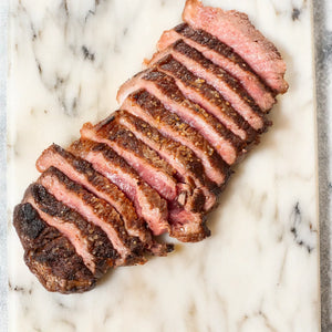 Perfectly seared and sliced USDA Prime Heritage Grass-Fed New York Strip steak arranged on a white marble board, showcasing a juicy, medium-rare interior and crisp, caramelized edges.