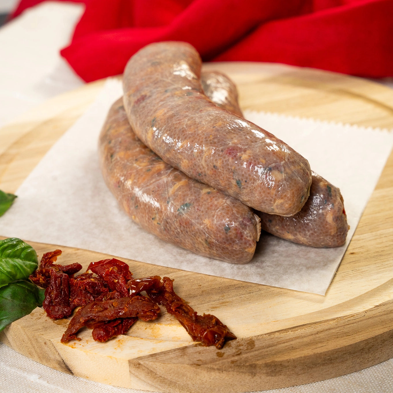 Raw All-Natural Joyce Farms Naked Chicken Sundried Tomato and Basil sausages rest, stacked on wax paper, on top of a wooden cutting board with basil leaves and dried red pepper resting on the side. A red hand-towel rests in the background.