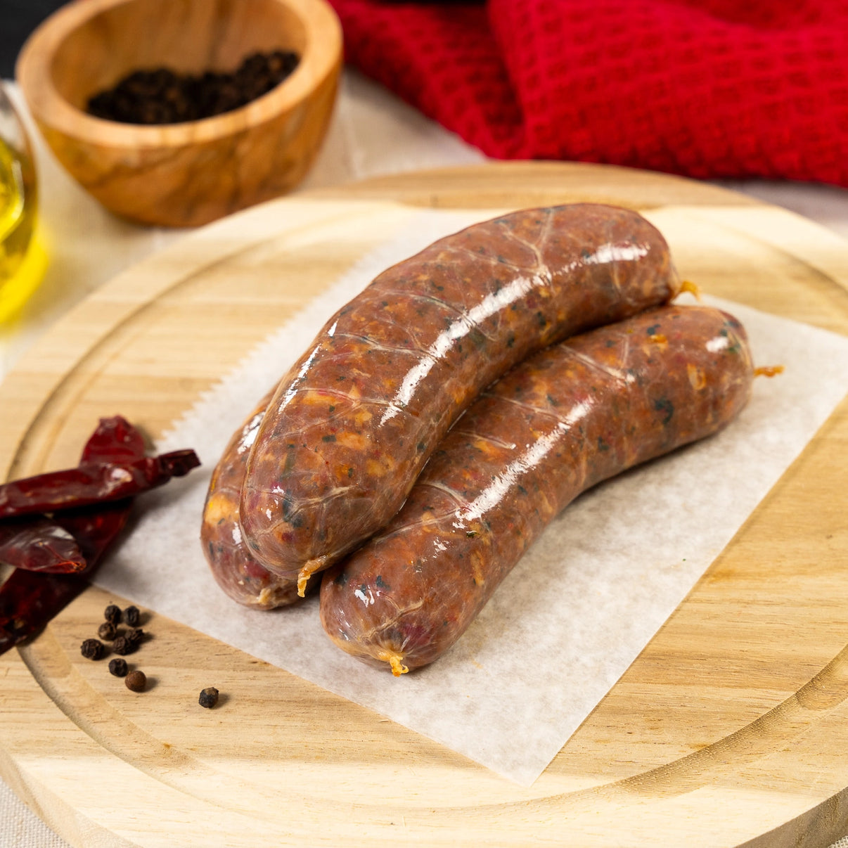 Joyce Farms all-natural, no antibiotic or growth hormone, naked chicken sausage chipotle flavor rests on wax paper on top of a wooden cutting board. Framing the sausages are dried peppers, peppercorns, a bottle of olive oil, a wooden ramekin of peppercorn, and a red hand-towel.