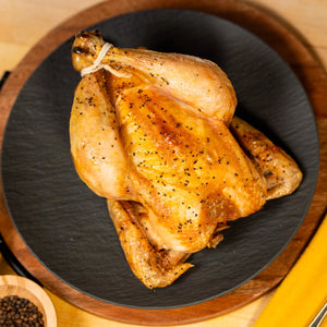 A cooked and trussed Joyce Farms Heritage Poulet Rouge® whole bird rests on a black plate sitting on top of a brown wooden presentation plate. It illustrates the tenderness and crispiness of the bird's skin.