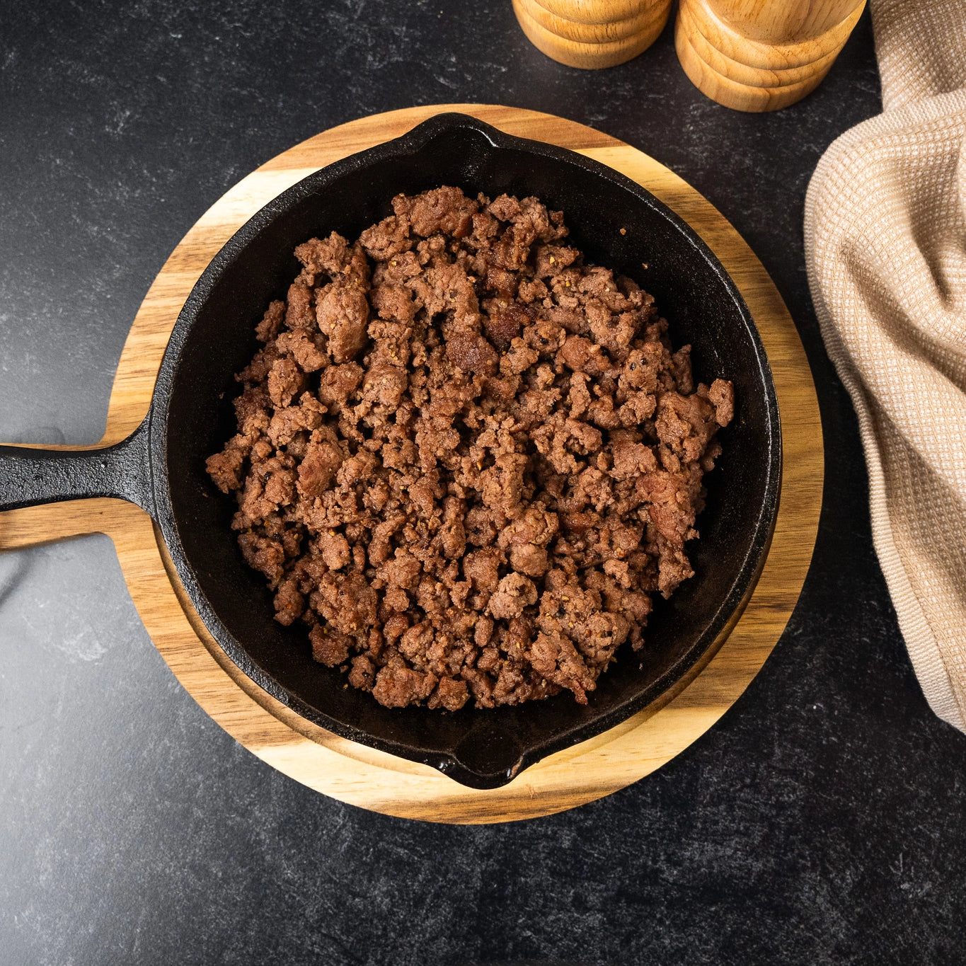 Cooked brown, Joyce Farms Heritage Aberdeen Angus ground meat rests in a cast iron skillet on a wooden trivet with a dark background.