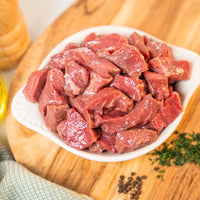 Raw heritage beef chunks in a white bowl on a wooden cutting board with herbs and spices.