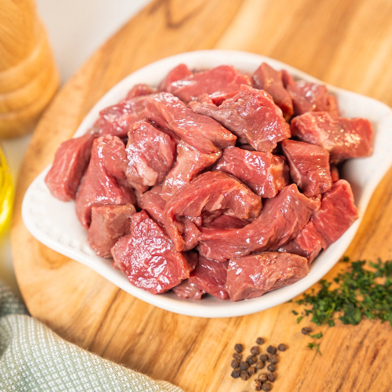 Raw heritage beef chunks in a white bowl on a wooden cutting board with herbs and spices.