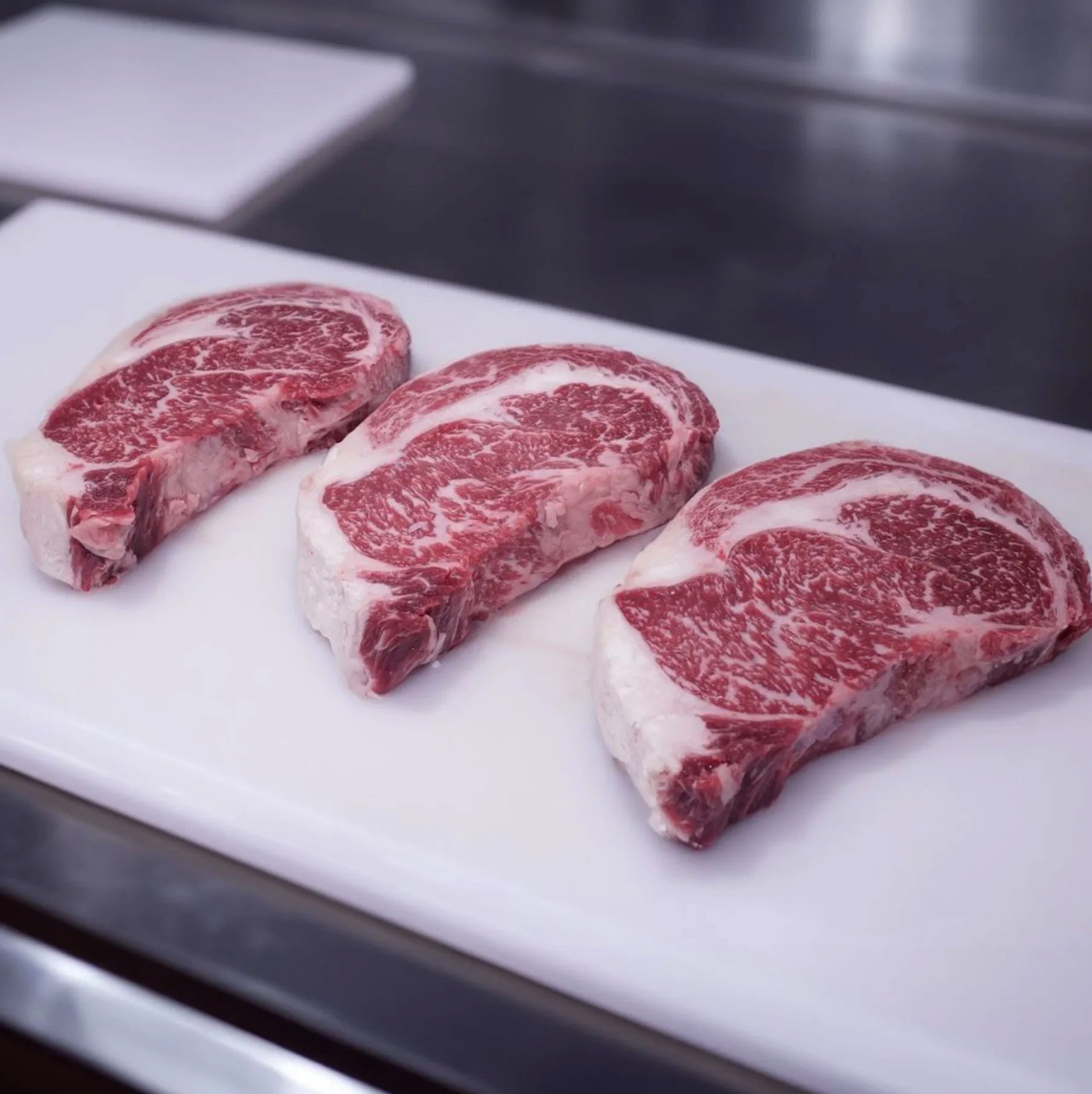 Joyce Farms 100% grass-fed and grass-finished Aberdeen Angus Heritage Beef, featuring three marbled ribeye steaks on a white cutting board.